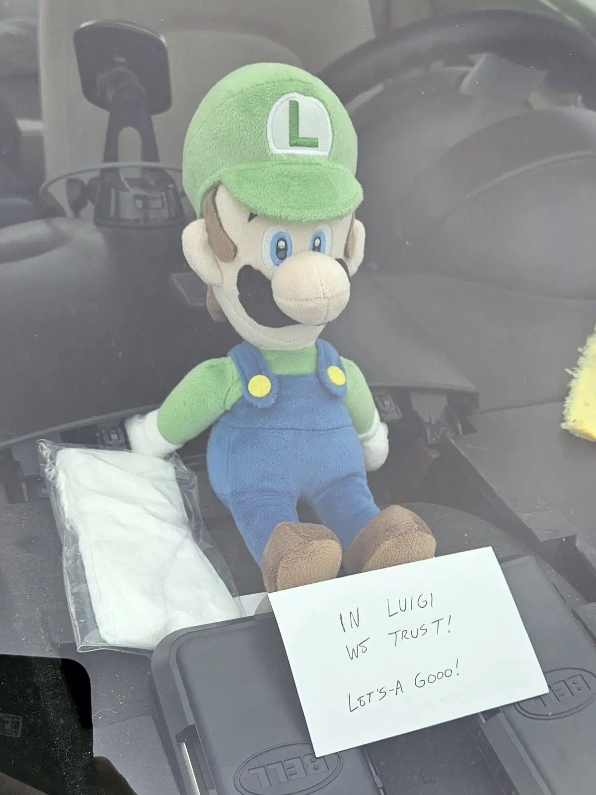 A plush Luigi doll sits in the driver's seat of a car, next to a handwritten note that reads 'IN LUIGI WE TRUST! LET'S-A…