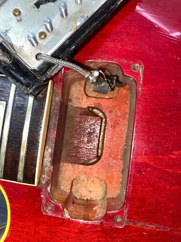 A close-up photo shows the underside of a red electric guitar's body, revealing a rusted metal pickup cavity with a broken…