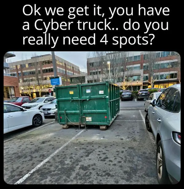 A large green dumpster is parked in the middle of a parking lot, occupying four marked parking spaces. Cars are parked on…