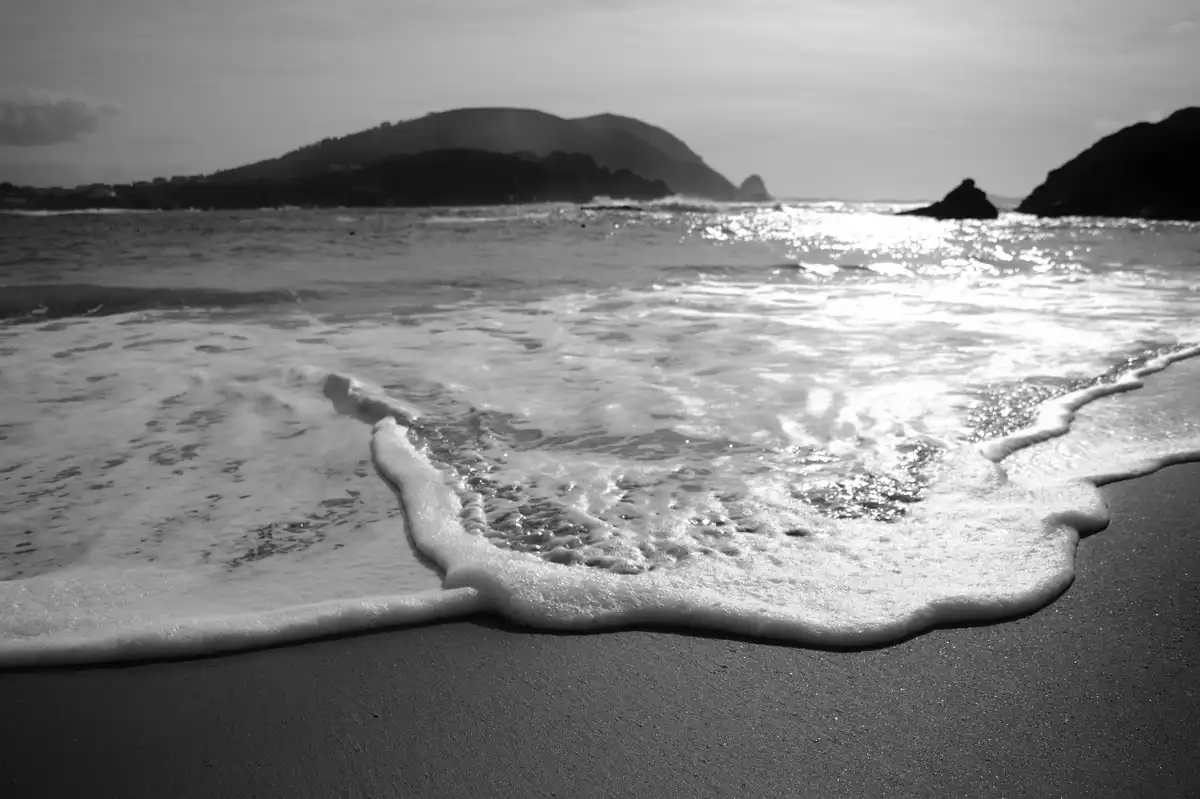 A black-and-white photo shows ocean waves gently rolling onto a sandy beach, with distant hills and rocks under a cloudy…