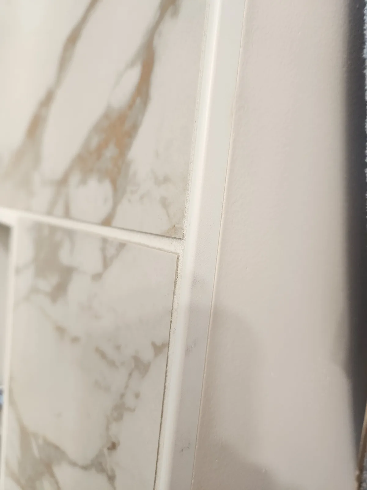 A close-up photo shows white marble-patterned tiles with visible grout lines, next to a plain white wall with a shadow. The…