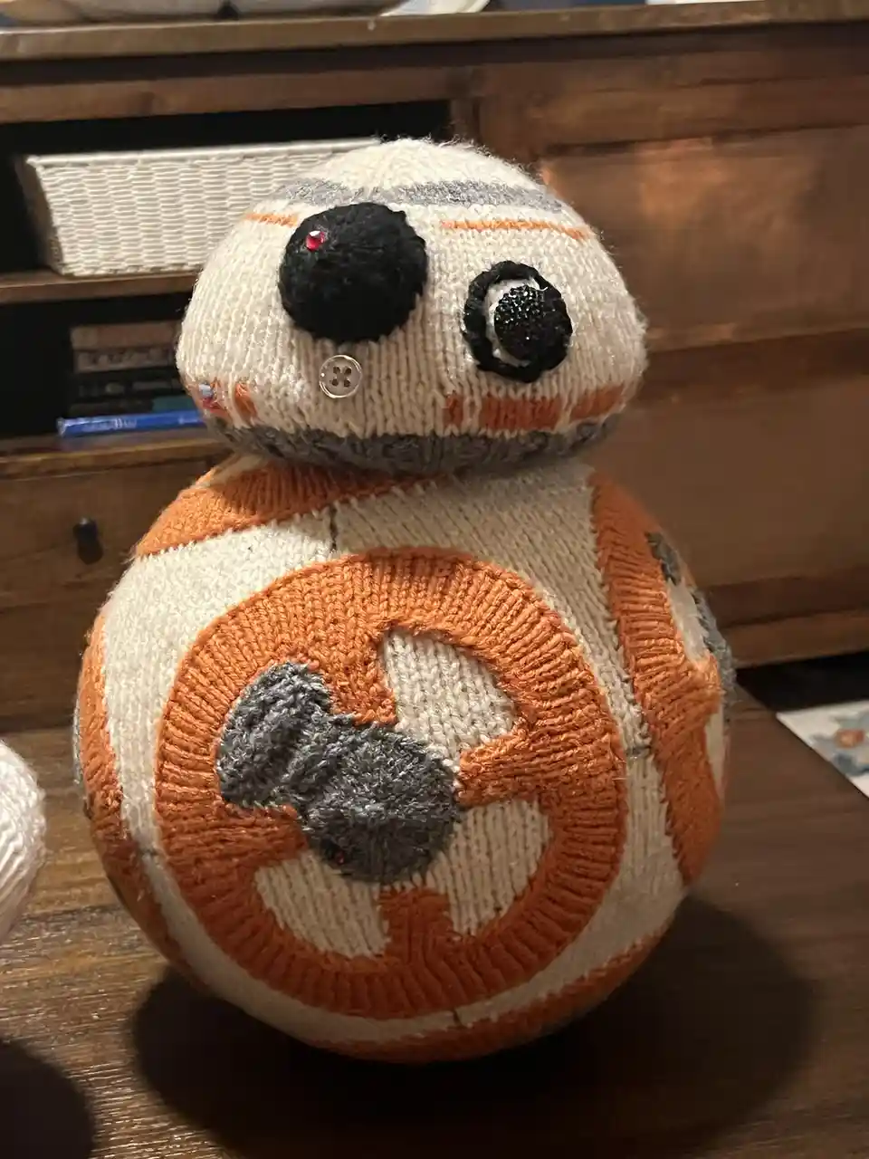 A handmade knitted plush toy resembling the Star Wars droid BB-8, featuring orange and white stripes, black button eyes with…