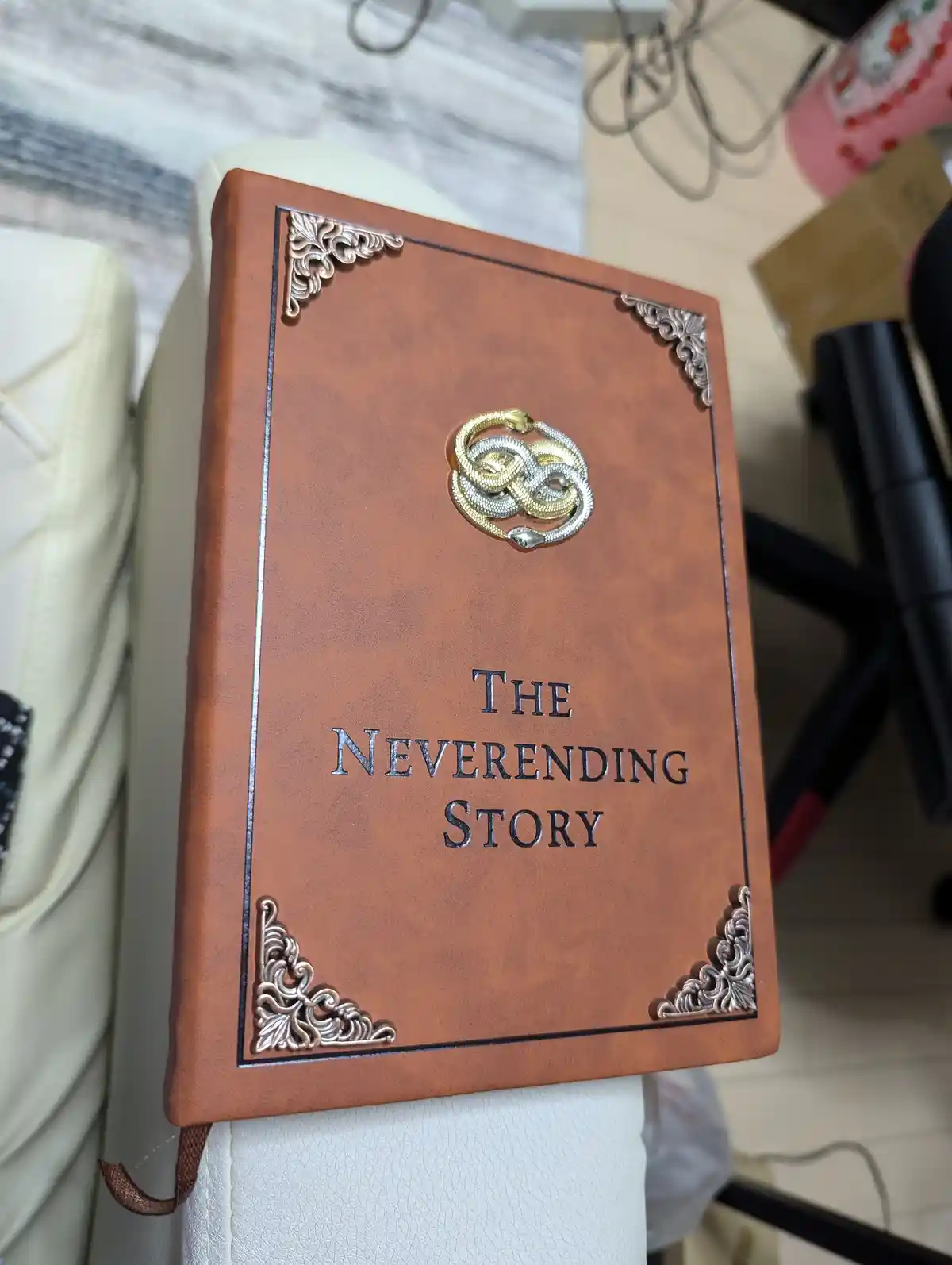 A brown leather-bound book with ornate silver corner embellishments and a central gold-and-silver intertwined snake emblem…