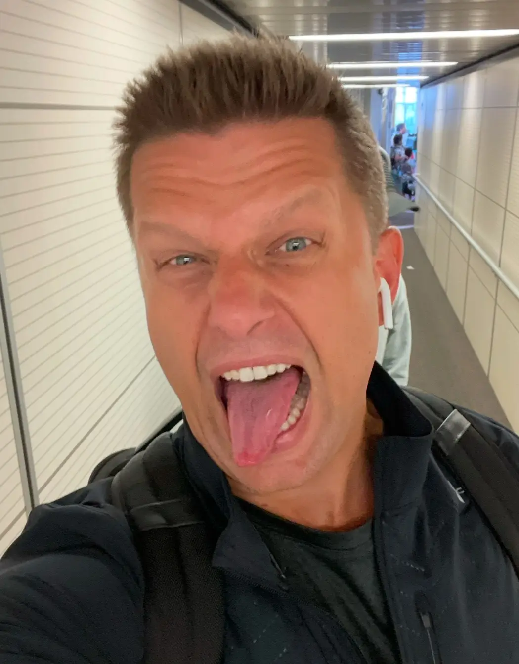 A man with short brown hair and blue eyes takes a selfie in a brightly lit indoor corridor, wearing a black jacket and white…