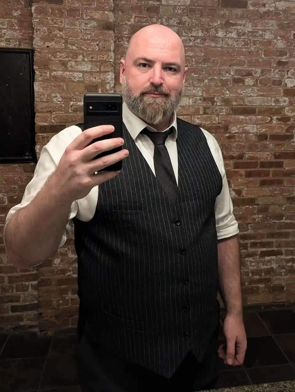 A bald man with a gray beard takes a selfie in front of a brick wall, wearing a white dress shirt, black tie, and a black…