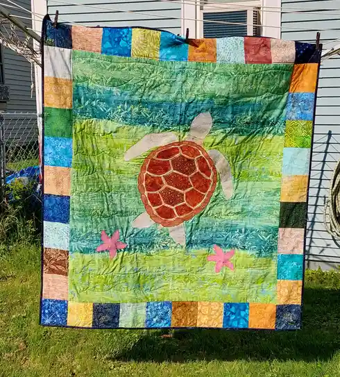 A colorful handmade quilt with a central sea turtle design and starfish motifs hangs outdoors on a clothesline, framed by a…