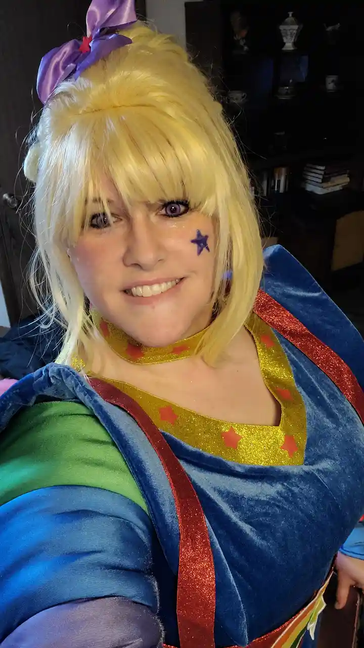 A person in a colorful cosplay costume with blonde wig, purple bow, star face paint, and glittery outfit poses with a slight…