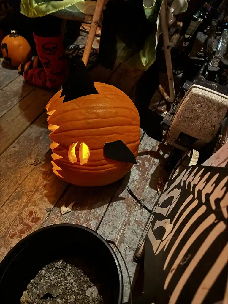 A carved Halloween pumpkin with glowing eyes and black paper bat wings sits on a wooden floor next to a black bucket and a…