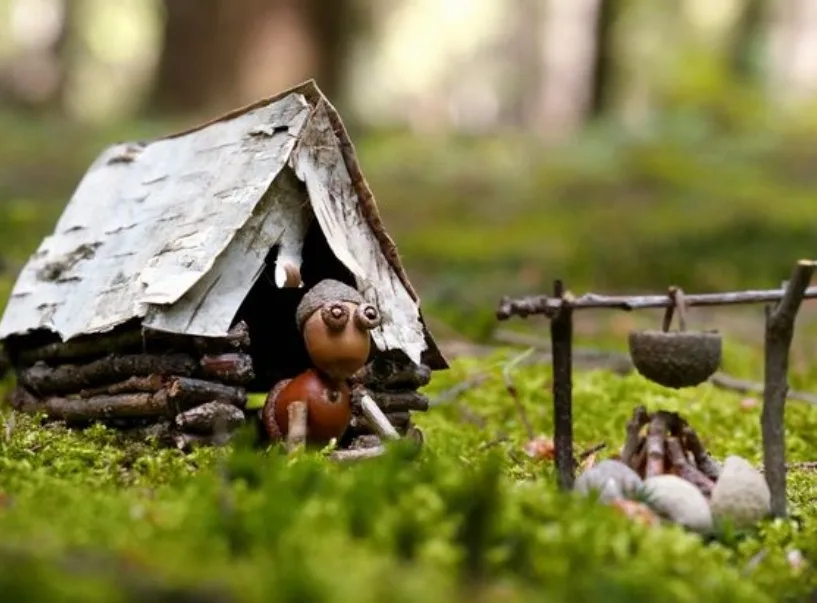 A tiny handmade figurine with large eyes sits in a miniature birch-bark hut in a mossy forest, next to a campfire and a…