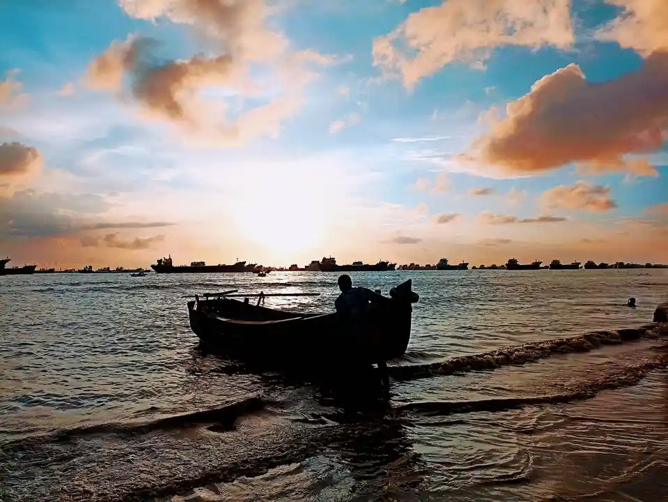 A silhouette of a person in a small boat at a beach during sunset, with a line of large cargo ships visible in the distance…