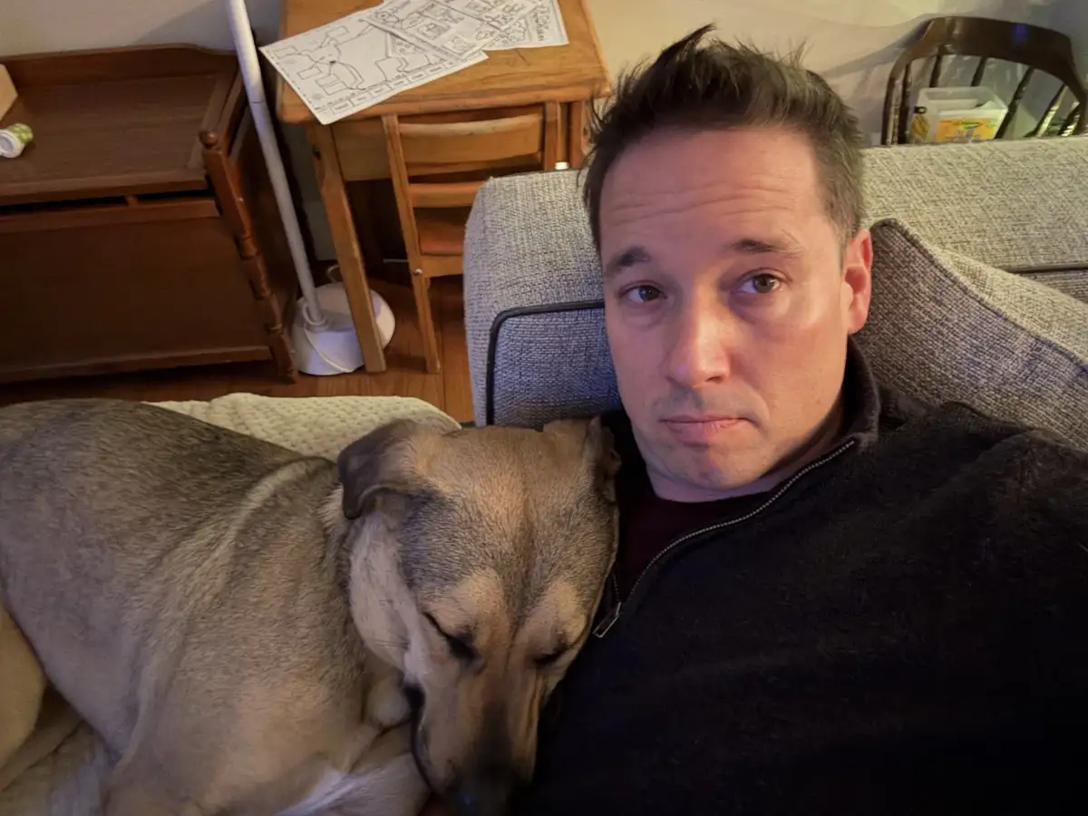A man in a dark zip-up sweater sits on a gray couch, looking directly at the camera with a weary expression, while a tan and…