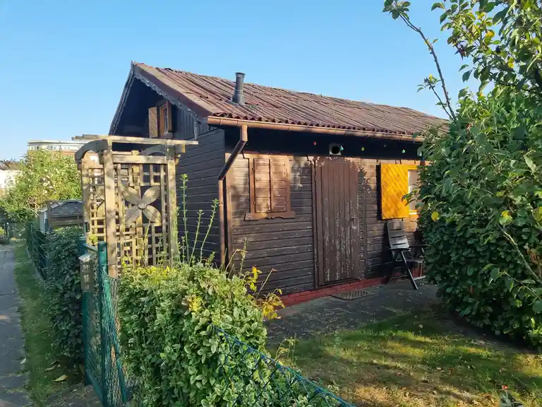 a small wooden cabin with dark brown siding, a rusty corrugated metal roof, and a single window with shutters, surrounded by…