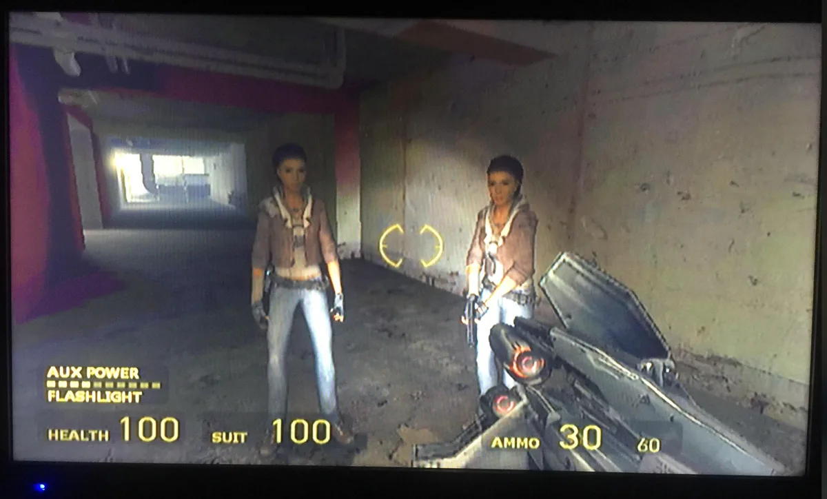 A first-person shooter game screenshot shows two identical female characters standing in a dimly lit, concrete corridor. The…