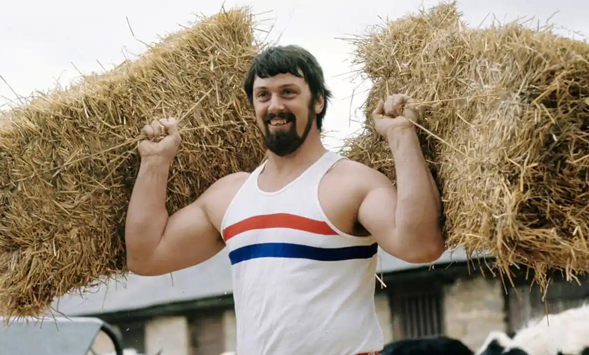 a muscular man with a beard wearing a white tank top with red and blue stripes stands outdoors holding two large hay bales…
