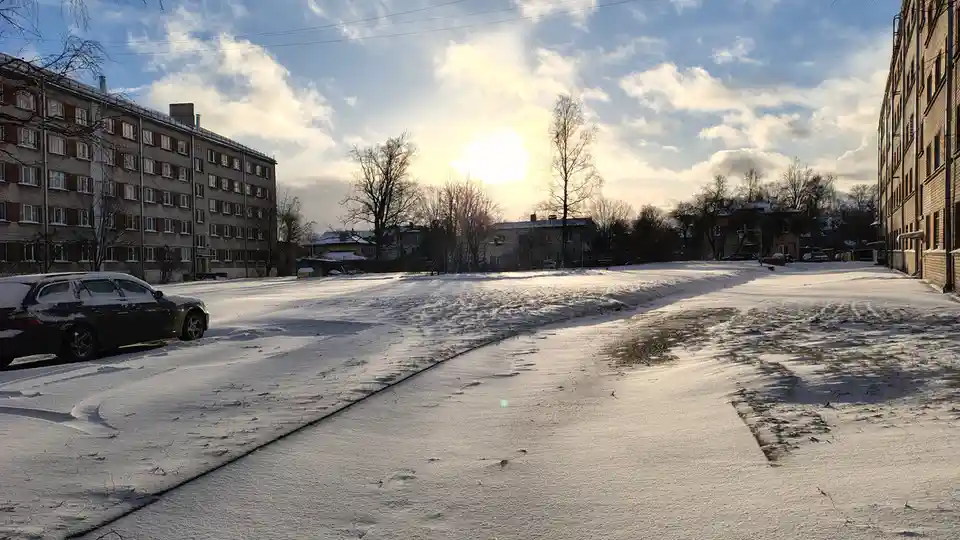 A snowy urban street with apartment buildings on either side and a dark car parked on the left, under a bright sun partially…