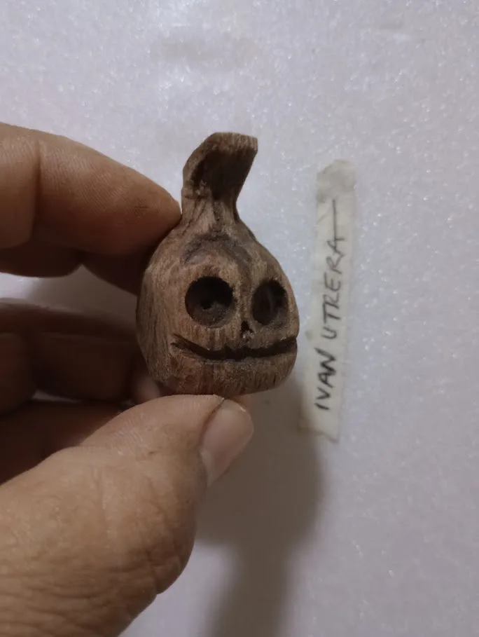 A hand holds a small, carved wooden object resembling a skull with large eye sockets and a wide grin, set against a white…