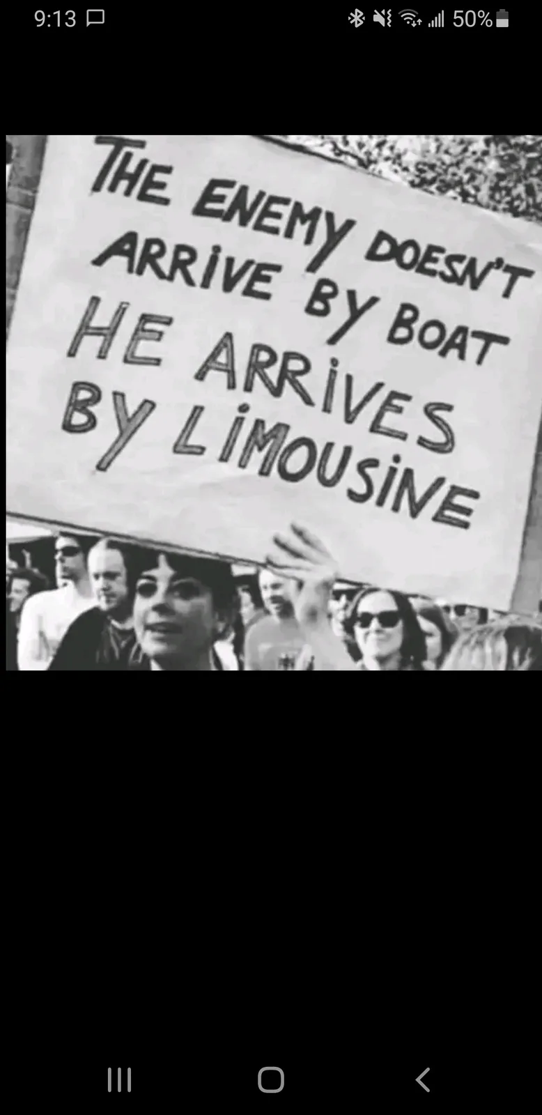 A black-and-white photo shows a crowd at a protest, with a woman prominently holding a handmade sign. The sign, written in…