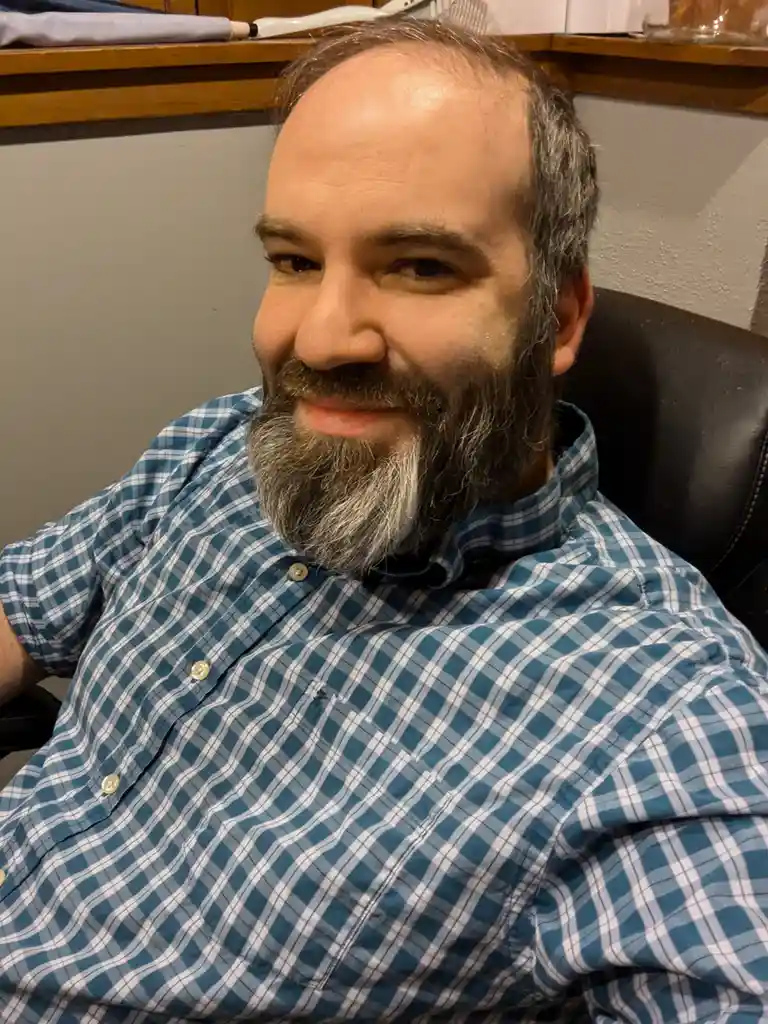 A middle-aged man with a graying beard and receding hairline, wearing a blue and white plaid shirt, sits in a chair and…