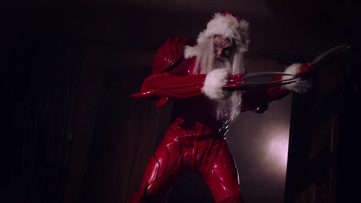 A person in a shiny red, muscular Santa Claus costume with a white beard and fur-trimmed hat is posed aggressively, holding…