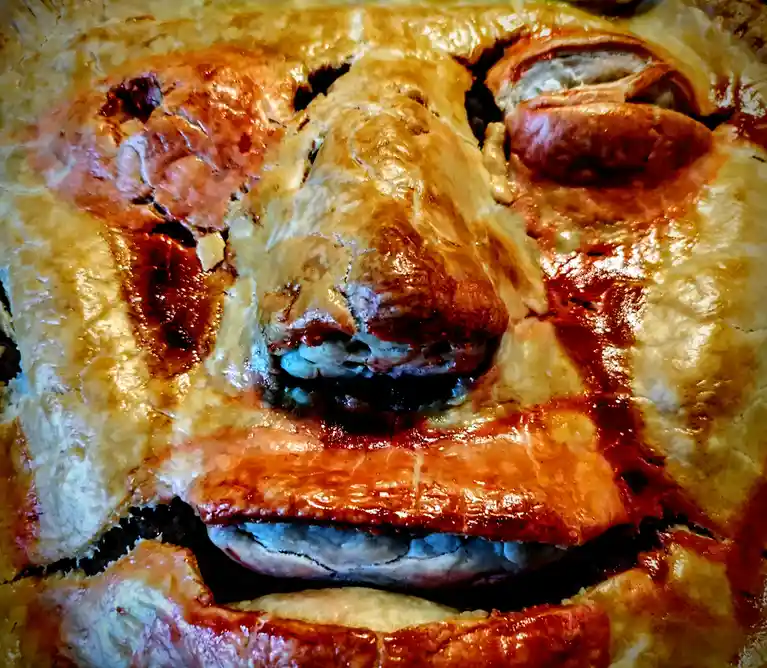A close-up of a golden-brown baked pie with a crust that has been shaped and baked to resemble a human face, complete with a…