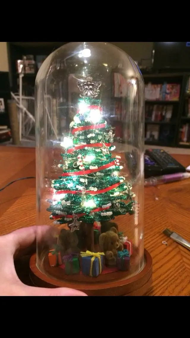 A hand holds a glass-domed display containing a miniature, illuminated Christmas tree decorated with red ribbons, white…
