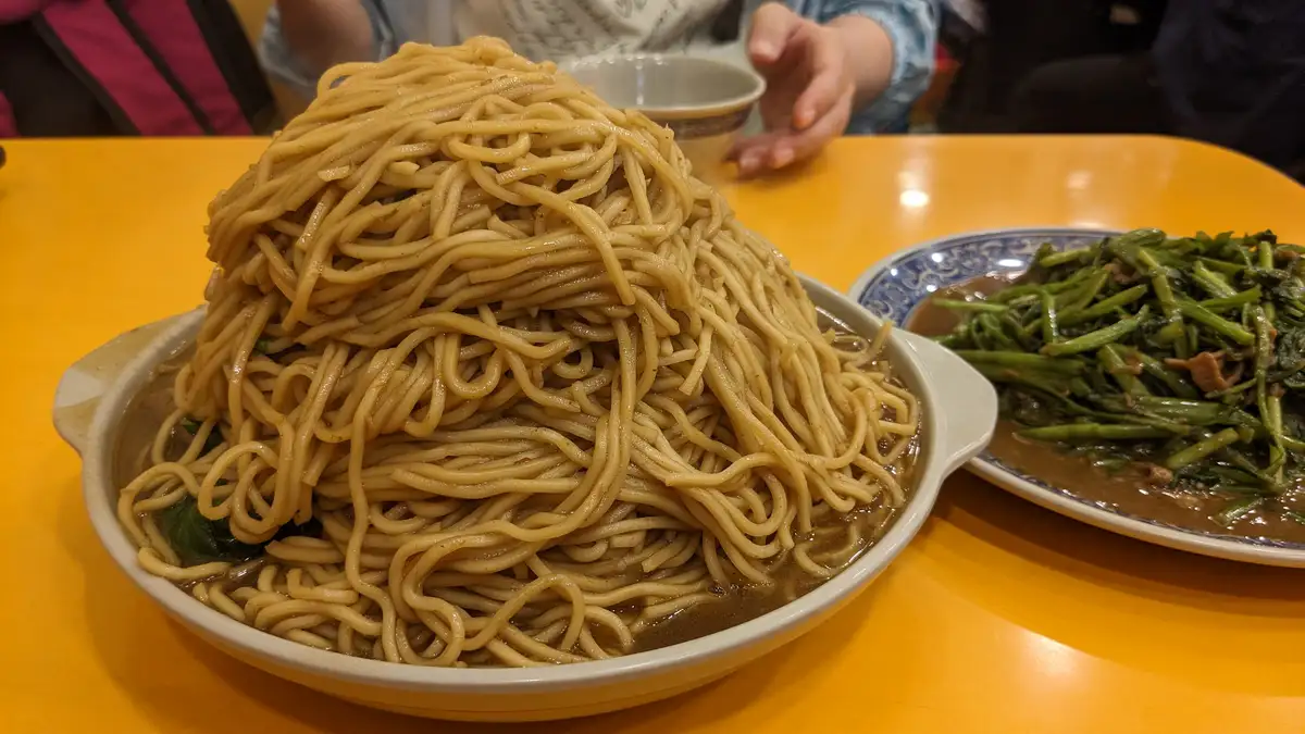 A massive, towering pile of noodles sits in a white bowl on a yellow table, with a side dish of greens nearby. A person's…
