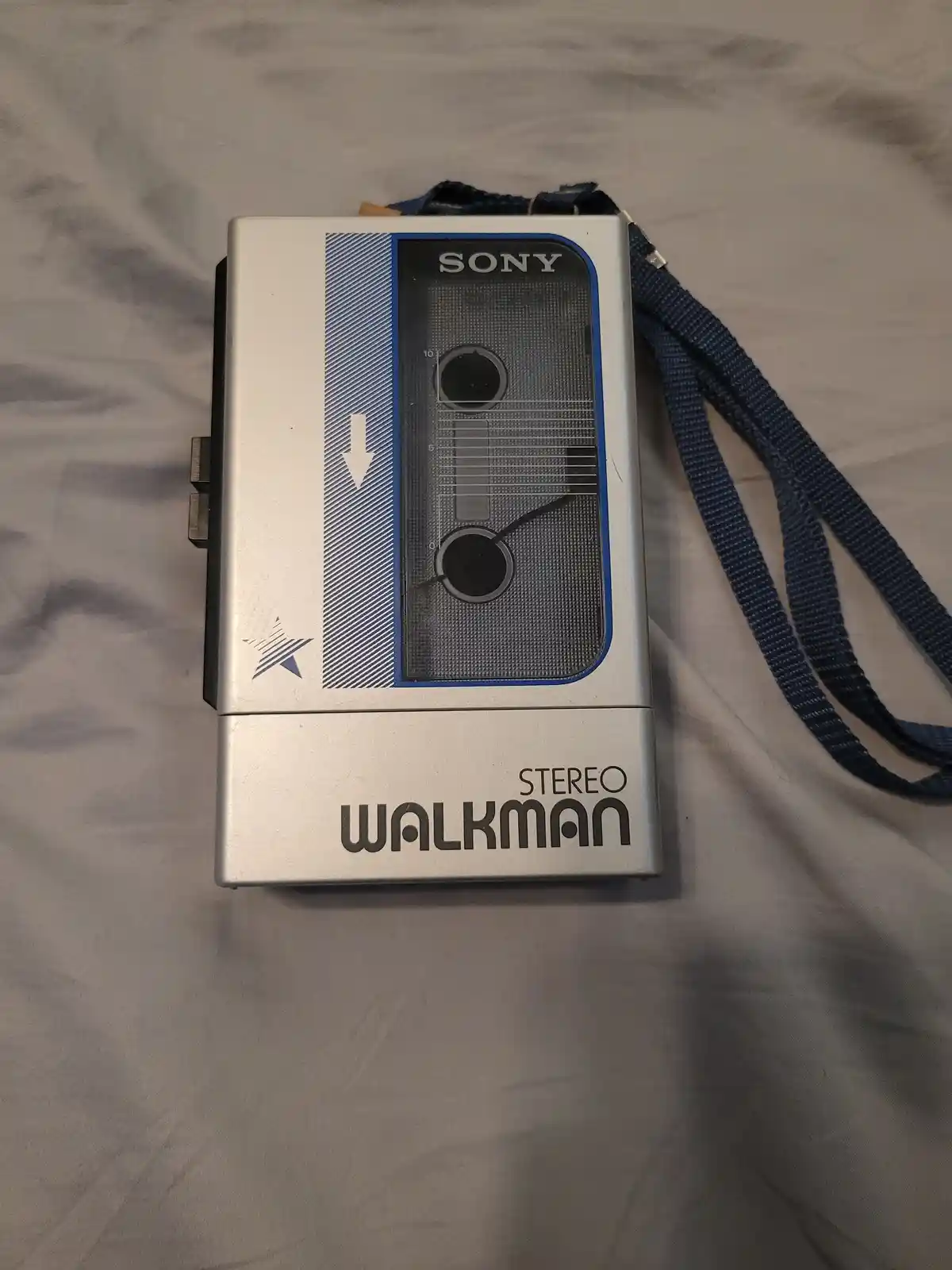 A silver Sony Stereo Walkman portable cassette player with a blue strap lies on a wrinkled gray fabric surface. The device…
