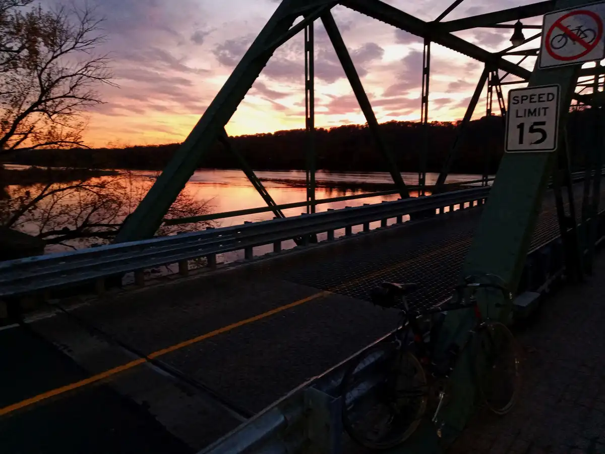 A bicycle is leaning against a bridge support next to a 'no bikes' sign and a 'speed limit 15' sign, with a beautiful sunset…