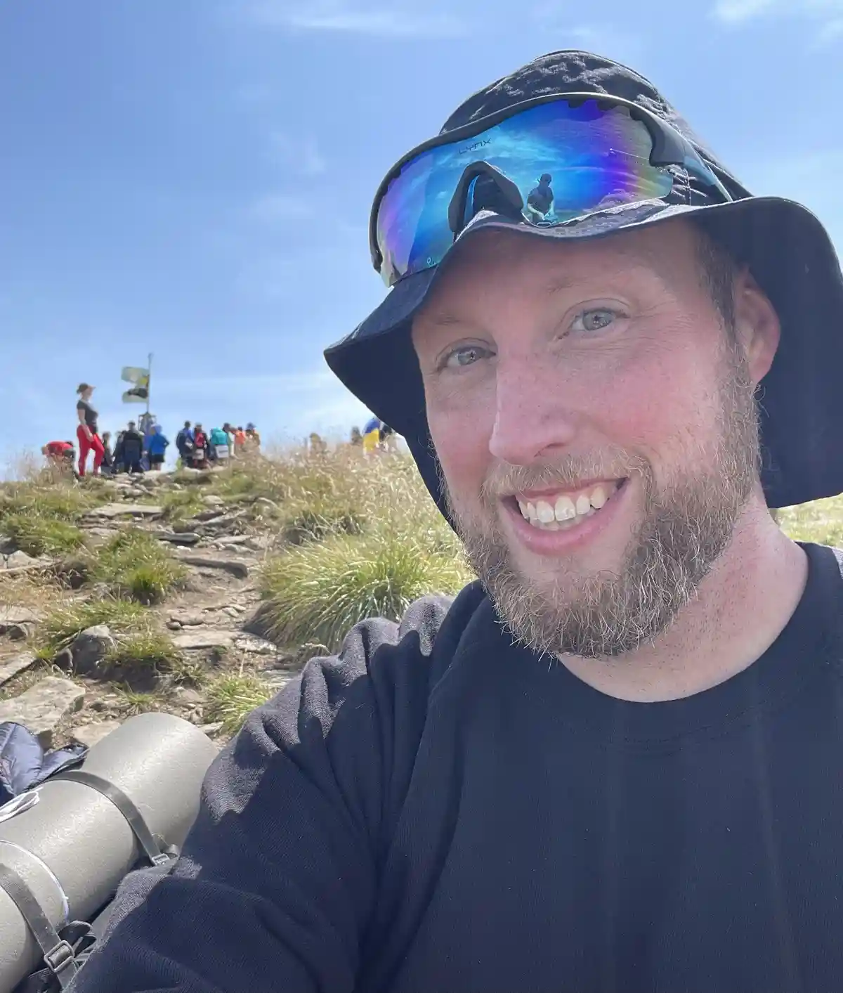 A man with a beard and a black bucket hat takes a selfie at the top of a mountain, smiling broadly. His reflective…