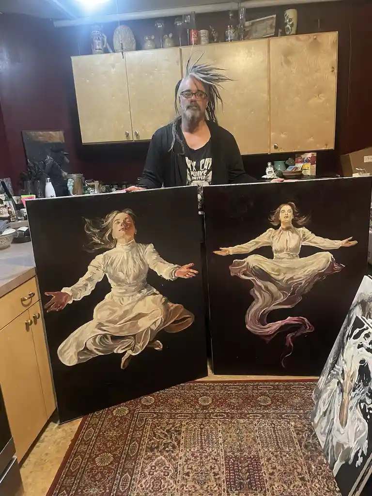 A man with dreadlocks and glasses stands in a kitchen holding two large paintings side-by-side. Both paintings depict a…