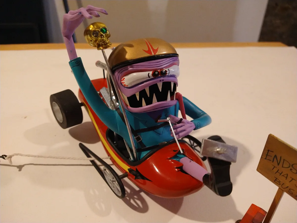 A custom toy monster truck driver with purple skin, sharp teeth, and a gold helmet is seated in a red race car, holding a…