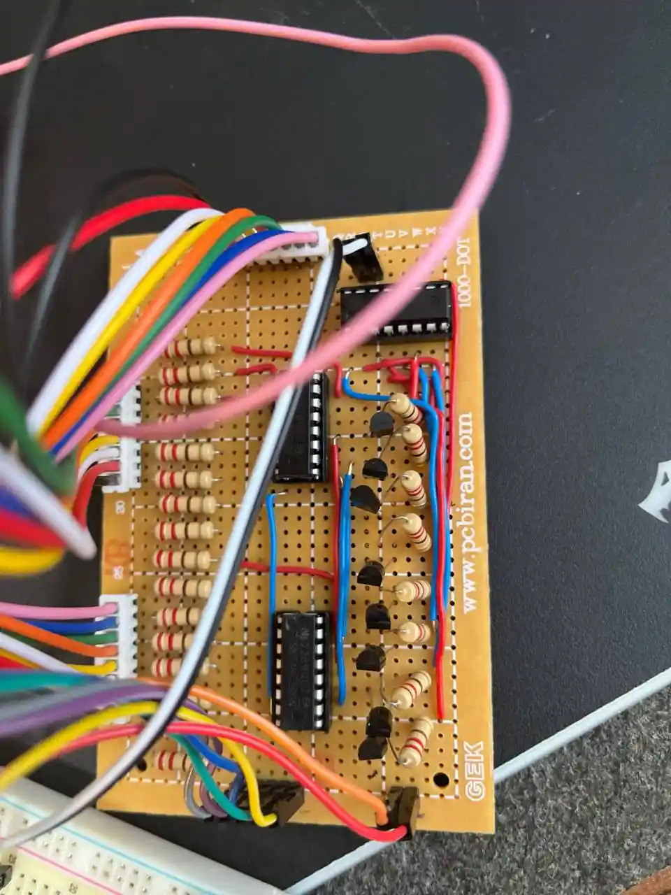 A close-up photo of a messy breadboard circuit with numerous colorful wires tangled around integrated circuits, resistors…