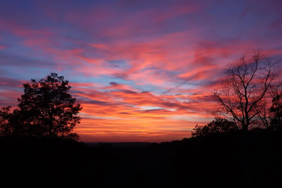 A vibrant sunset with fiery orange, red, and purple clouds over a dark silhouette of trees and hills. The overlaid text…