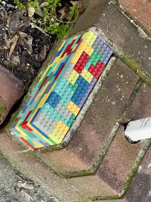 A colorful arrangement of Lego bricks forms a rainbow pattern on a weathered brick patio, surrounded by moss and fallen…