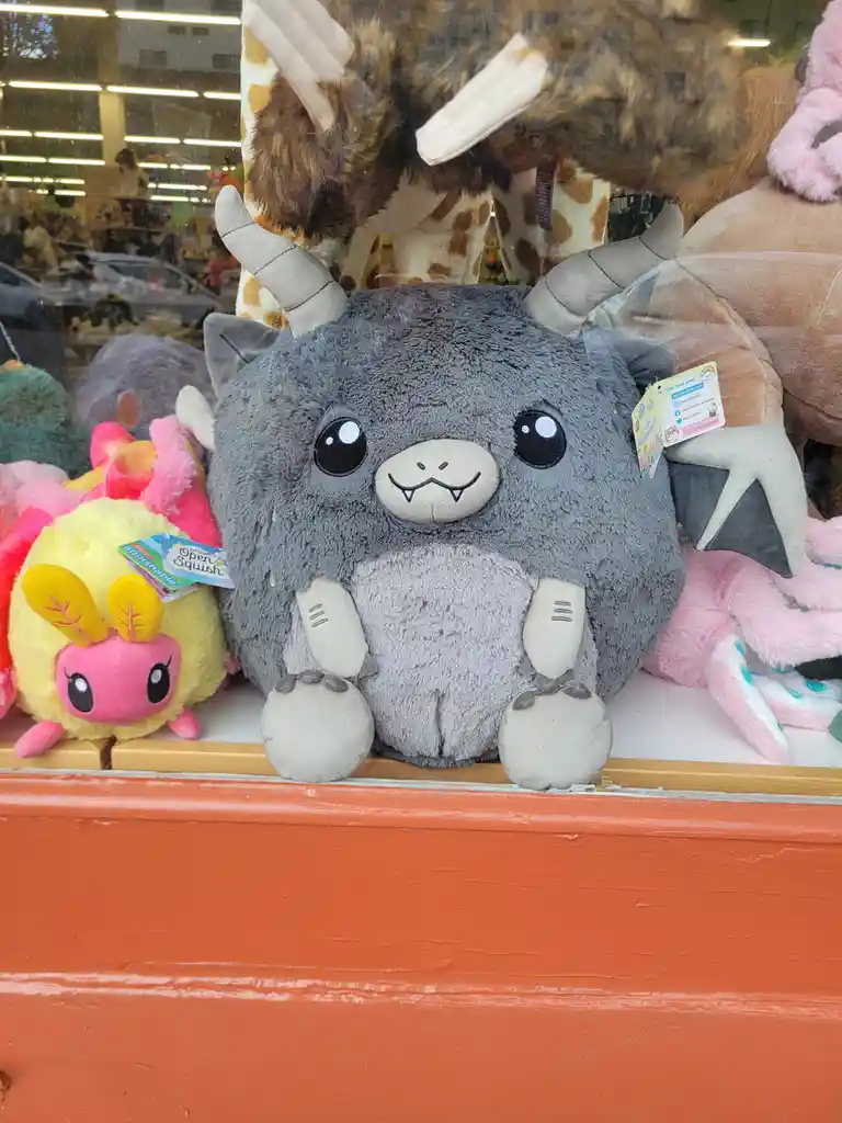 A grey, round, plush toy resembling a cute monster with small horns, fangs, and big eyes sits in a store window display…