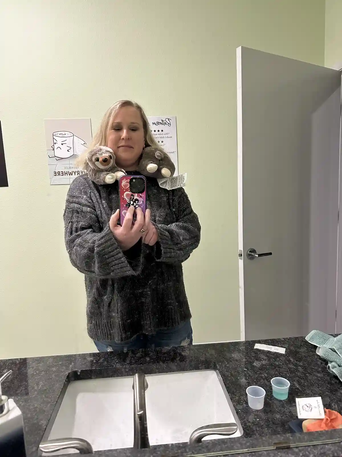A woman stands in a bathroom mirror selfie, holding a phone with a pink case, while two stuffed sloths are perched on her…