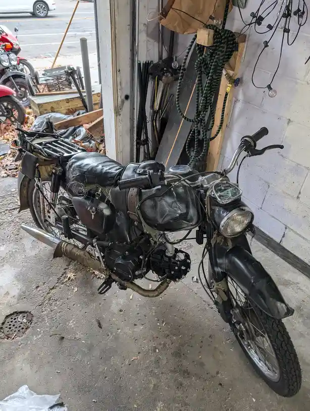 A heavily modified, rugged black motorcycle with a custom leather seat, exposed engine, and various straps and gear is…