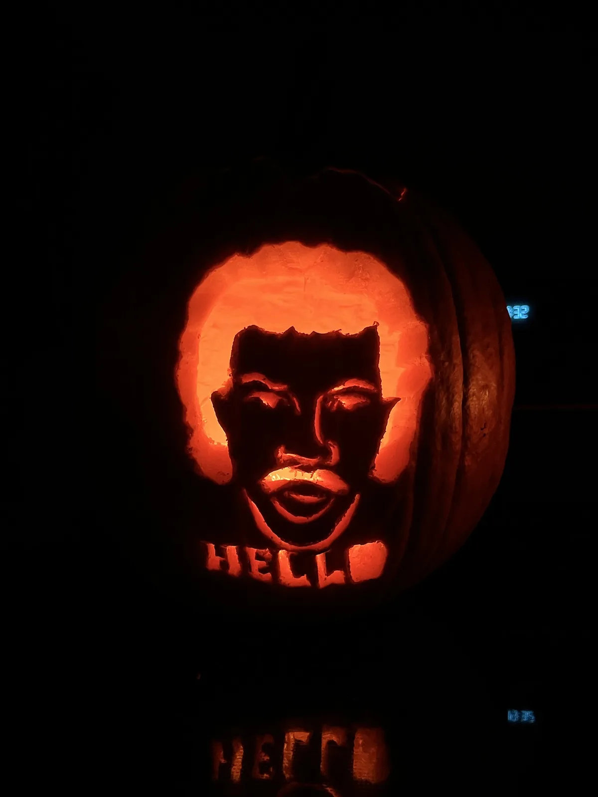 A carved pumpkin illuminated from within at night, featuring a detailed silhouette of a smiling man's face with short hair…