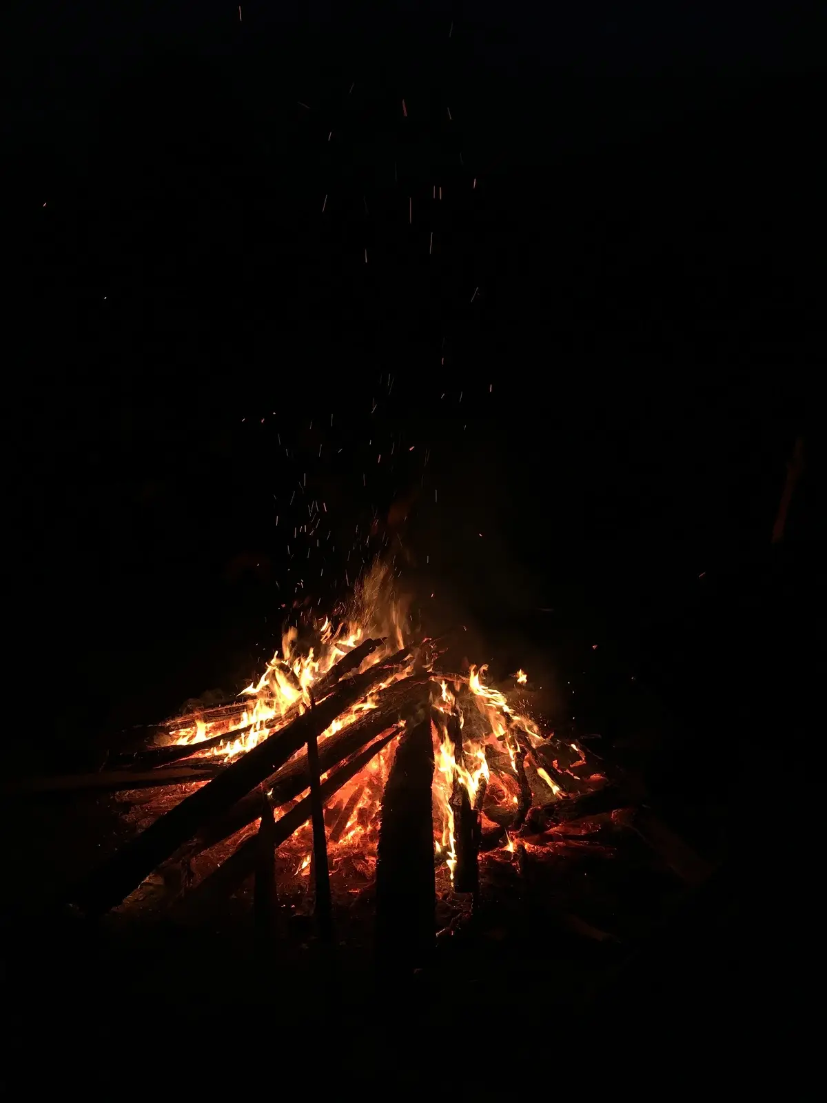 A nighttime photograph of a large, roaring campfire with bright orange flames and sparks flying upward against a completely…