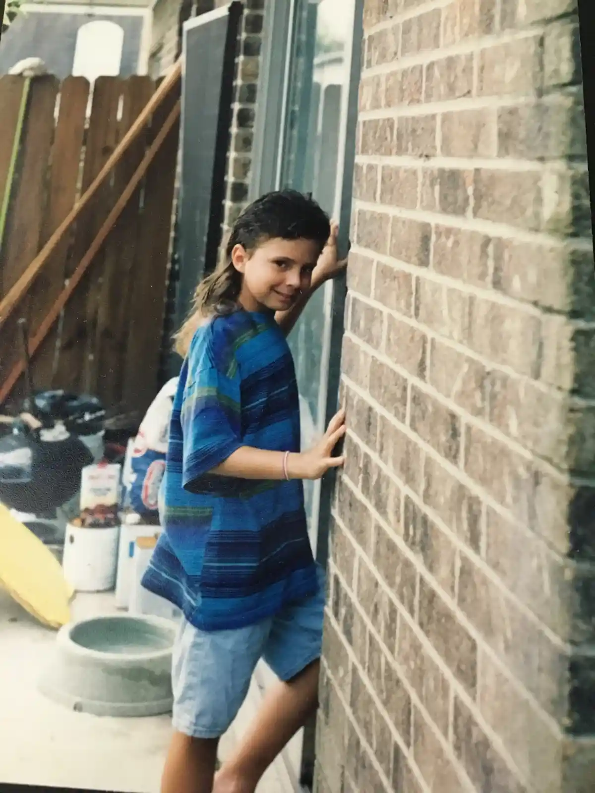 A young boy with long hair, wearing a blue striped shirt and denim shorts, leans against a brick wall in a backyard, smiling…