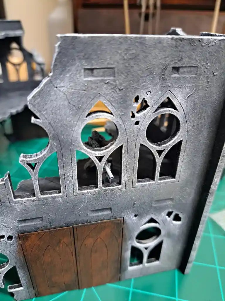 A close-up photo of a dark grey, gothic-style miniature building with ornate, pointed windows and a brown wooden door…