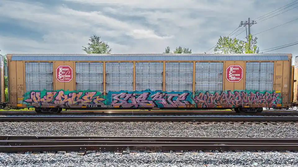 A Kansas City Southern Lines freight train car sits on tracks under a cloudy sky, its lower half covered in colorful…