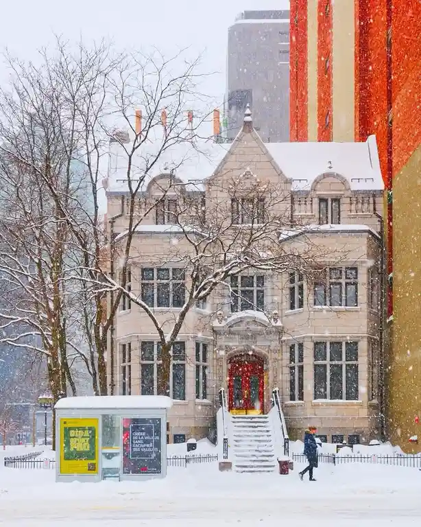 A snowy urban scene shows a person walking past a historic stone building with red doors, bare trees, and snow-covered…