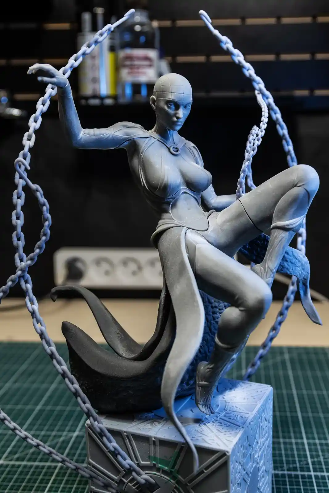 A gray, unpainted statue of a female figure with chains swirling around her, sitting atop a detailed base, is shown in a…