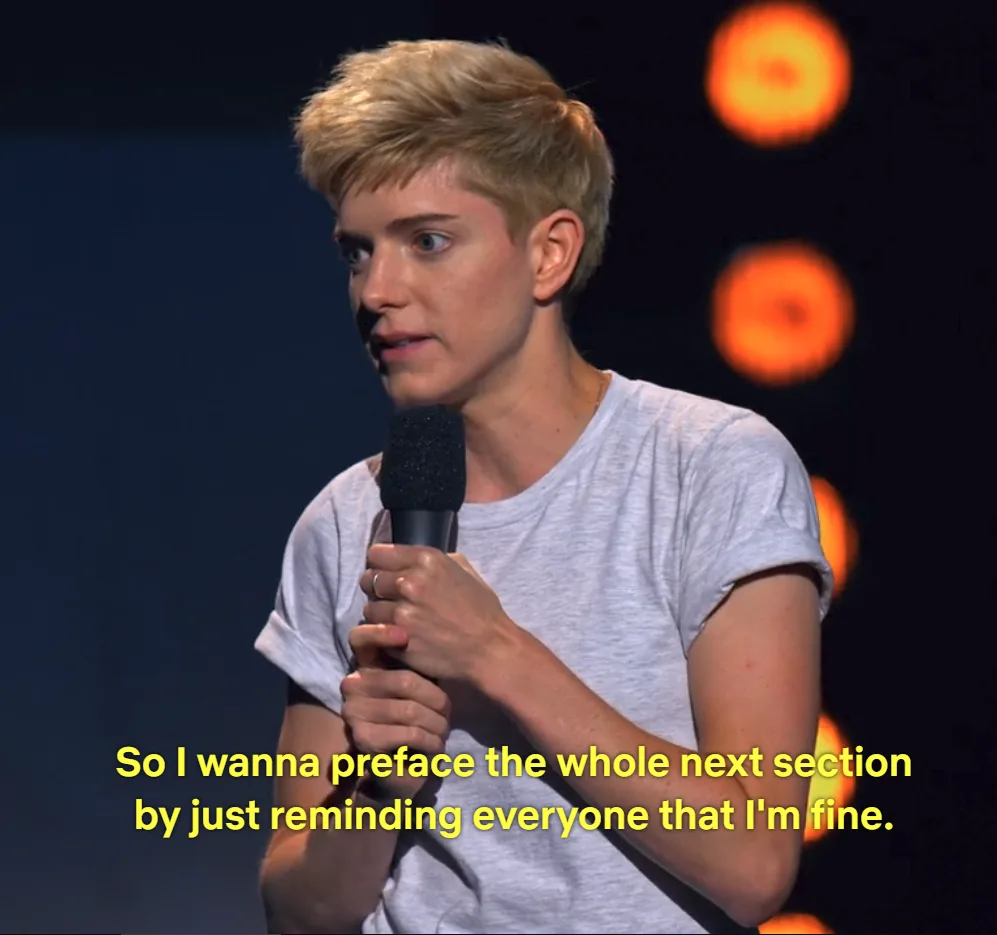 A person with short blonde hair, wearing a gray t-shirt, holds a microphone on a stage with blurred orange lights in the…