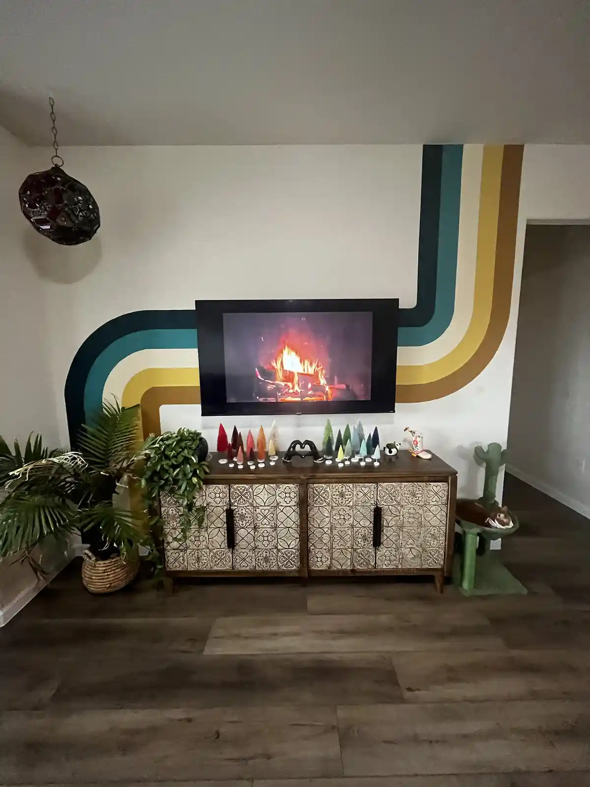 A living room features a TV mounted on a wall with retro-style curved stripes, displaying a realistic fireplace. Below the…