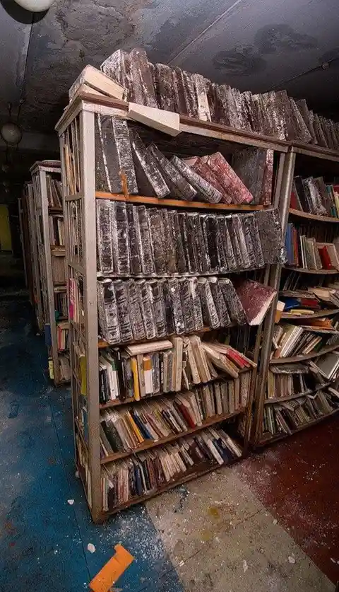A dimly lit, abandoned library with dusty, crumbling bookshelves overflowing with old, moldy books. The top shelves are…