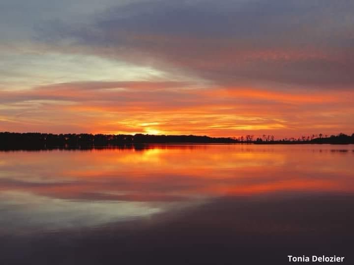 A serene sunset over a calm body of water, with vibrant orange and purple hues reflecting in the still surface. The horizon…