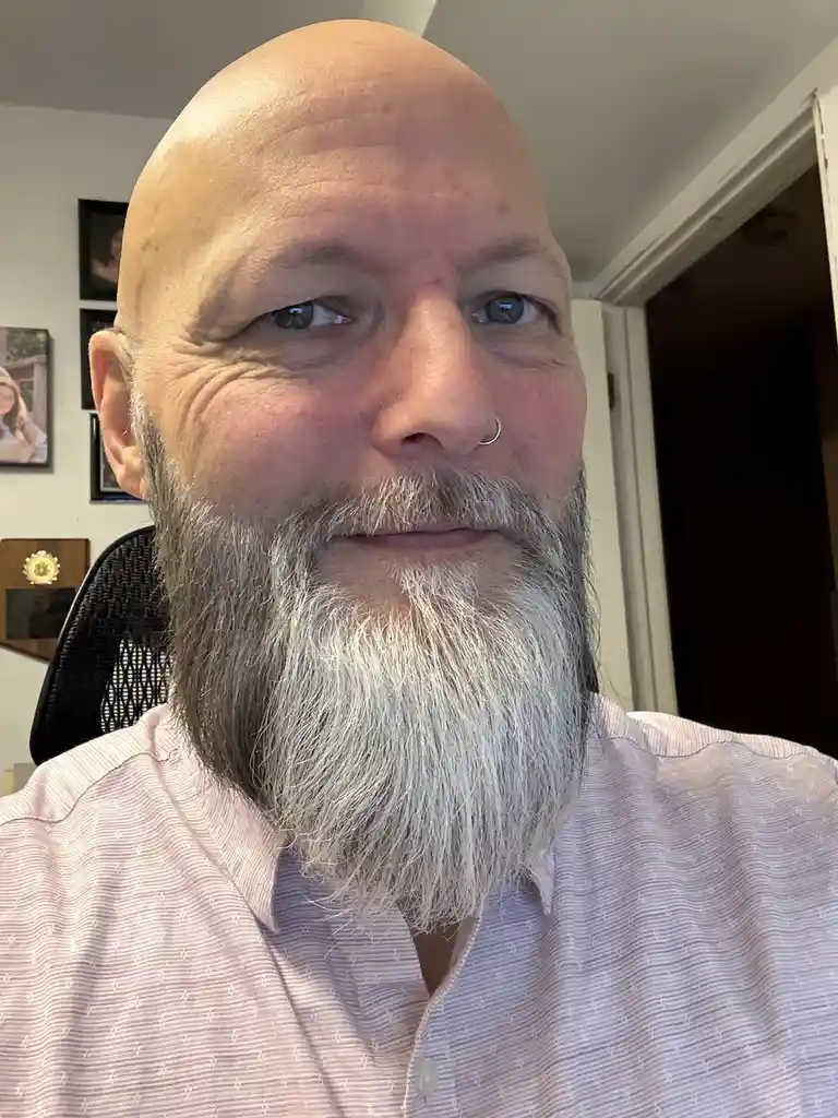 A close-up selfie of a bald man with a long, full gray beard and a nose ring, wearing a pink patterned shirt. He is looking…