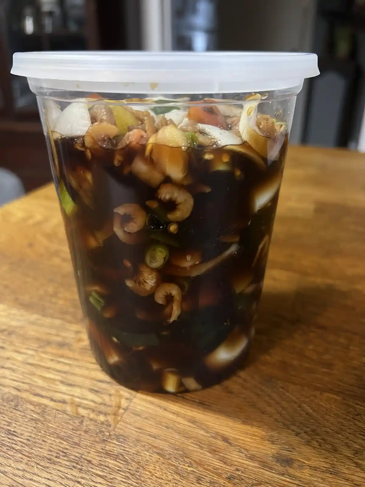 A clear plastic cup with a white lid sits on a wooden table, filled with a dark liquid and various ingredients including…